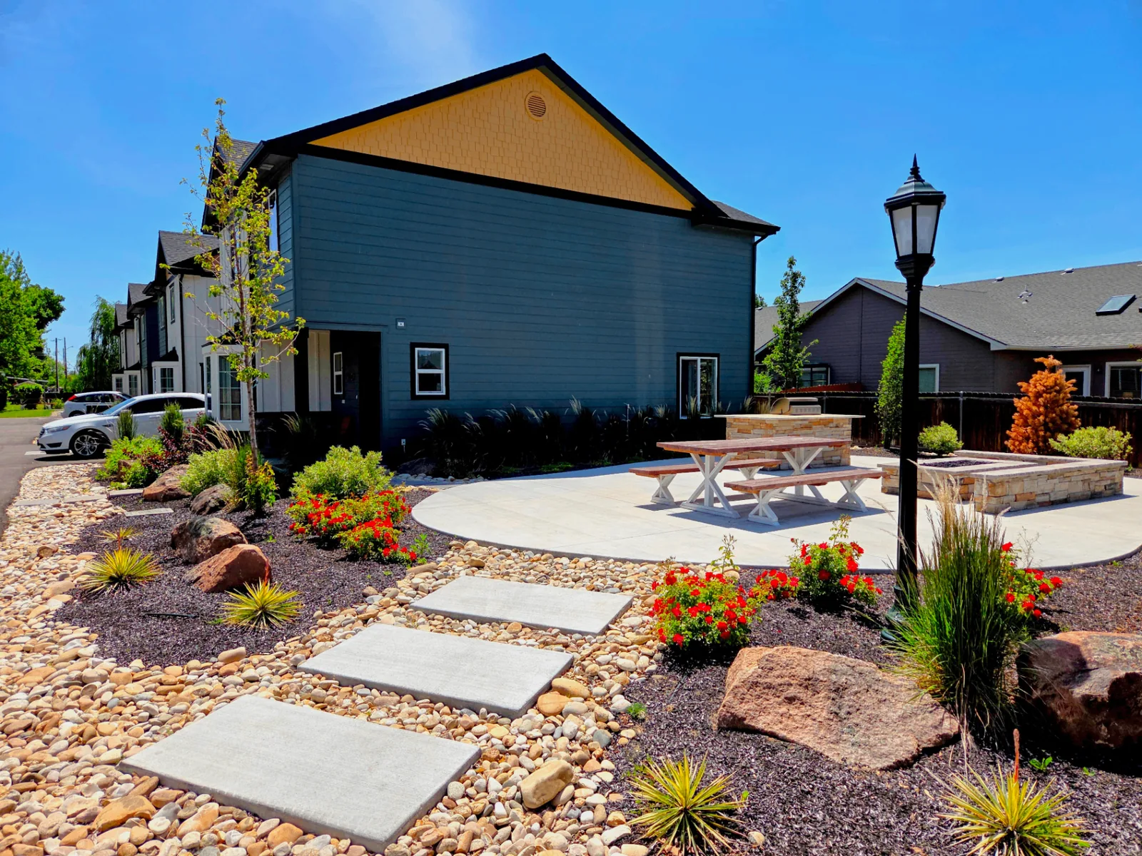Rising Sun Sober Living home exterior in Idaho with landscaped patio, picnic table, stone fire pit, and red flower beds under a blue sky